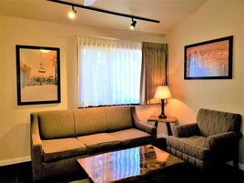 Sala de estar com sofá, poltrona e mesa de centro. Há uma janela com cortinas, um abajur em uma mesinha lateral e quadros nas paredes.