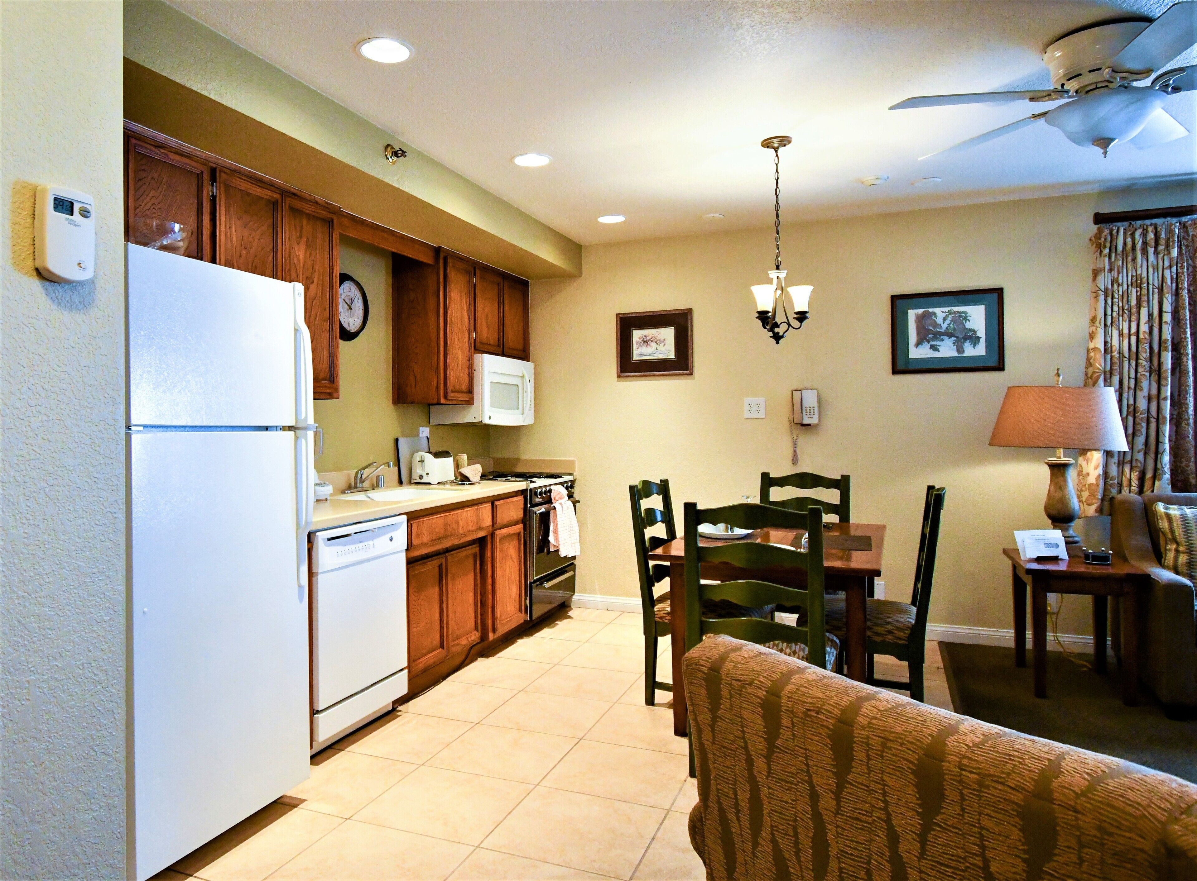 A compact kitchen with wooden cabinets, a microwave, and a refrigerator. There is a dining area with a table and chairs, and a ceiling fan above.
