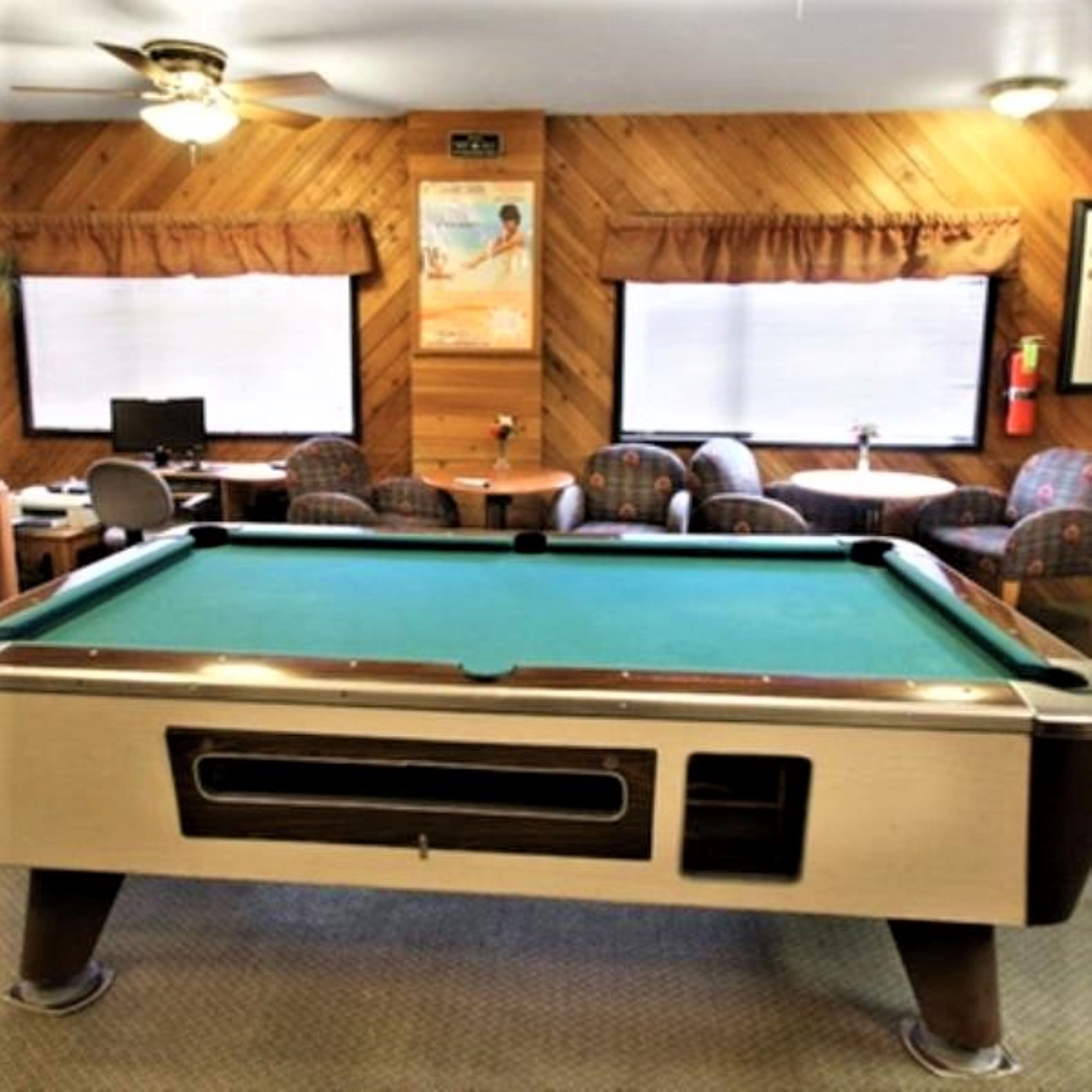 A pool table in a room with wooden walls, two windows, and a ceiling fan.