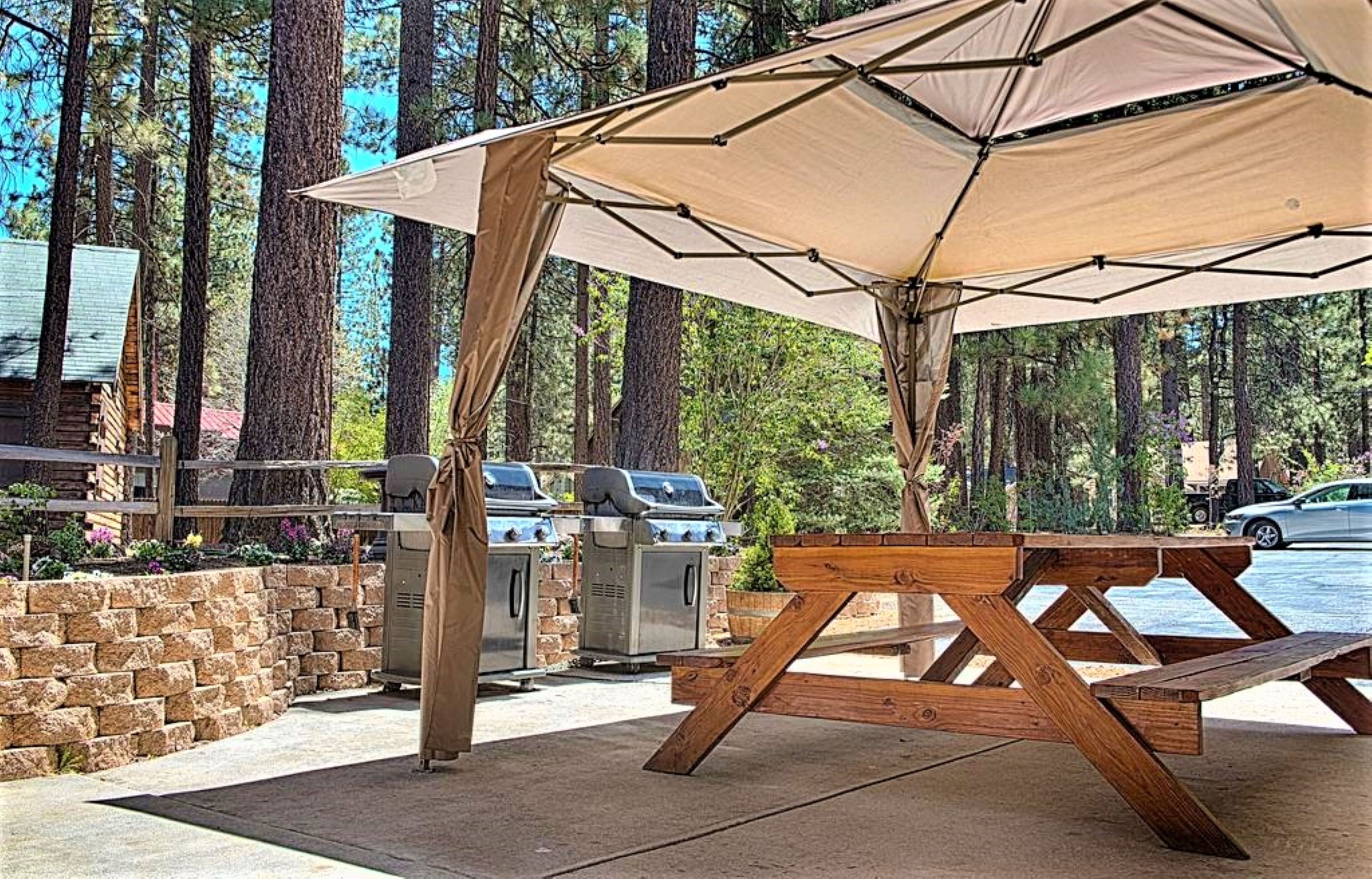 An outdoor patio with a wooden table and a beige canopy, surrounded by tall trees.