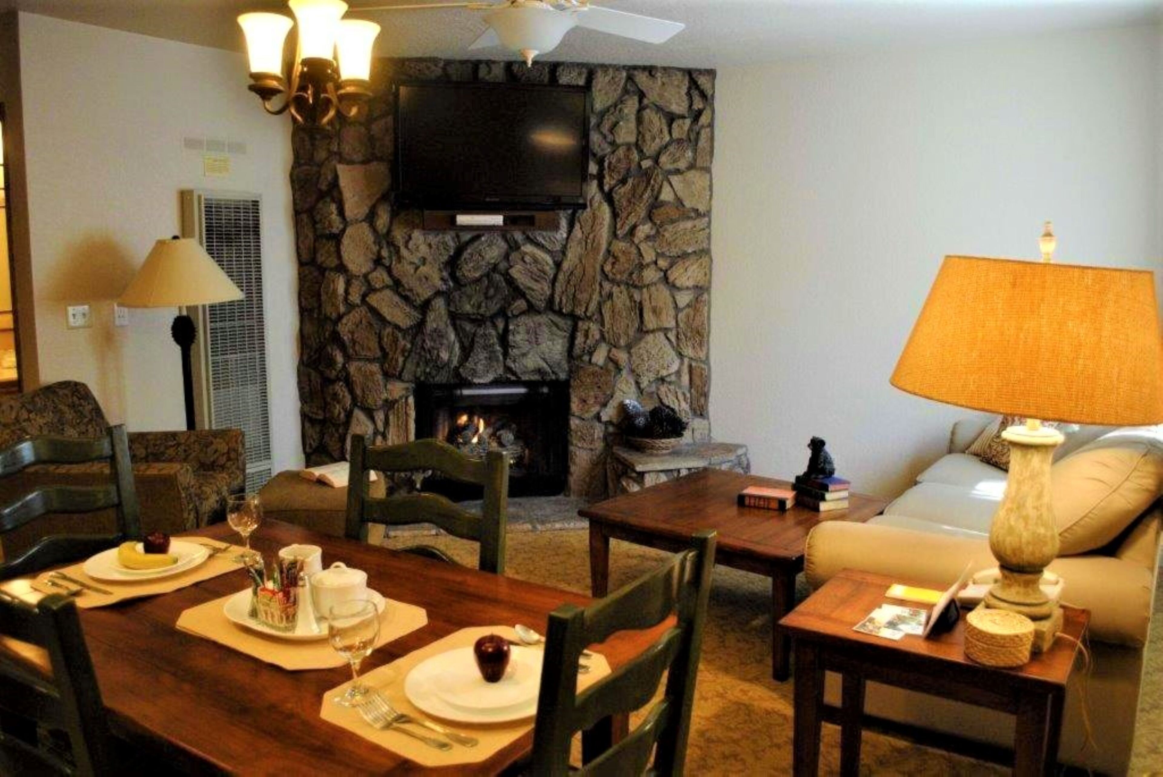 A living room with a stone wall, fireplace, flat-screen TV, dining table set for two, and a sofa.