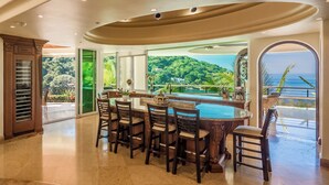 A spacious kitchen with a large island, a dining area with chairs, and a view of a lush landscape and ocean. - Luxury Beach Frontage Villa For Rent (Boca de Tomatlán)