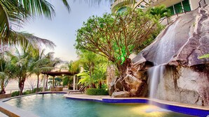 A pool with a waterfall, surrounded by palm trees and a stone wall. - Luxury Beach Frontage Villa For Rent (Boca de Tomatlán)
