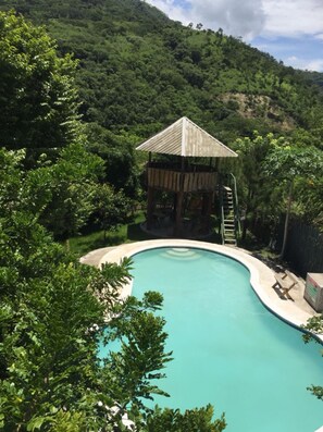 A swimming pool surrounded by lush greenery, a wooden structure with a roof, and a staircase leading up to it.