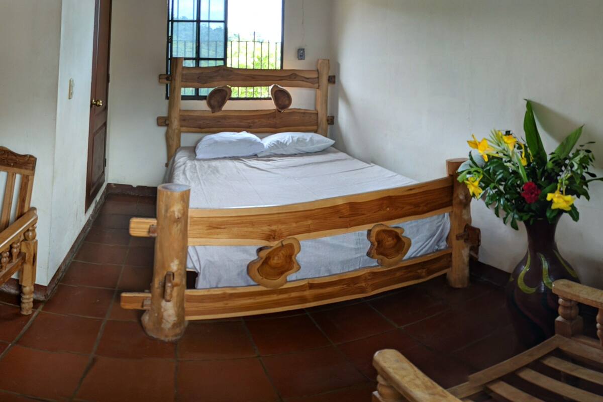 Una cama de madera con cabecera, una silla de madera, un banco de madera y un florero con flores.