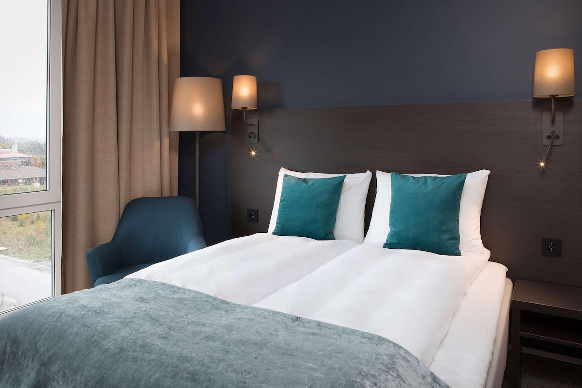 A hotel room with a large bed, two wall-mounted lamps, a chair, and a view through a window.
