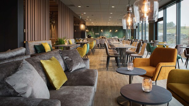 A modern restaurant with a mix of seating options including sofas and tables, large windows offering a view, and pendant lighting.