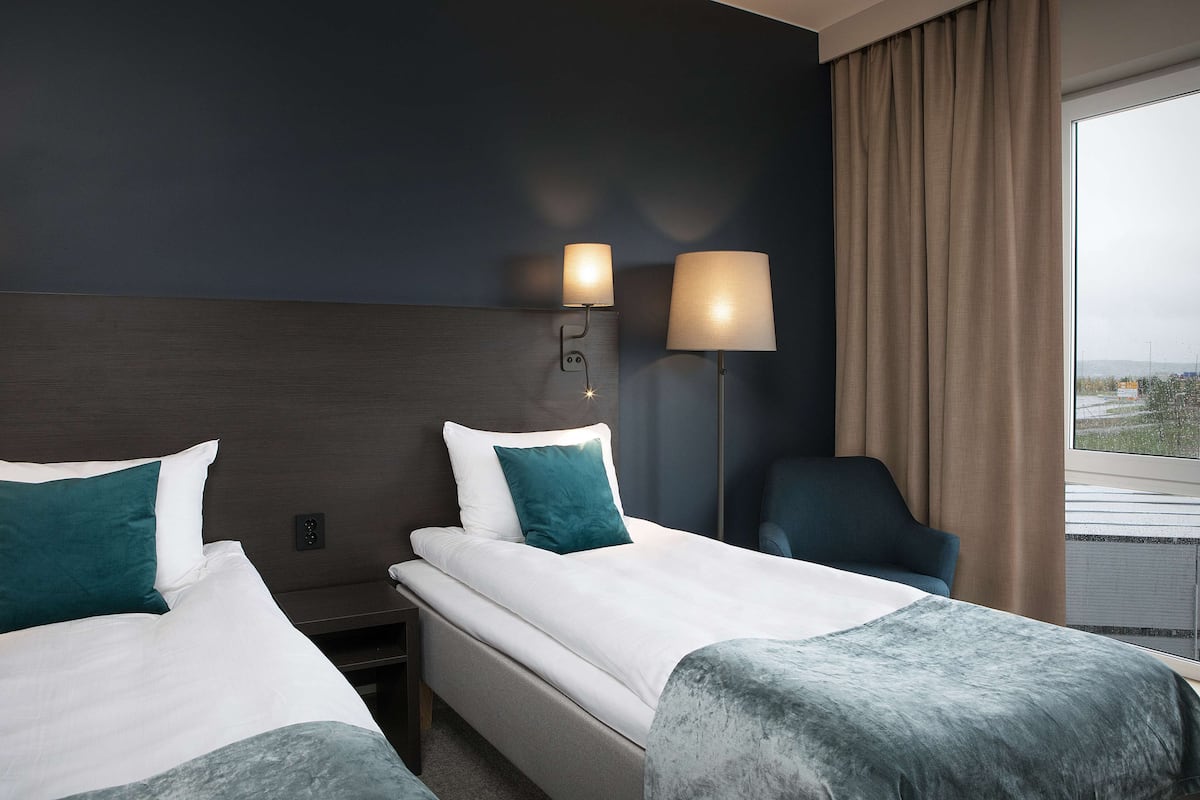 Habitación de hotel con dos camas, una mesita de noche, una lámpara, una silla y un ventanal con cortinas.