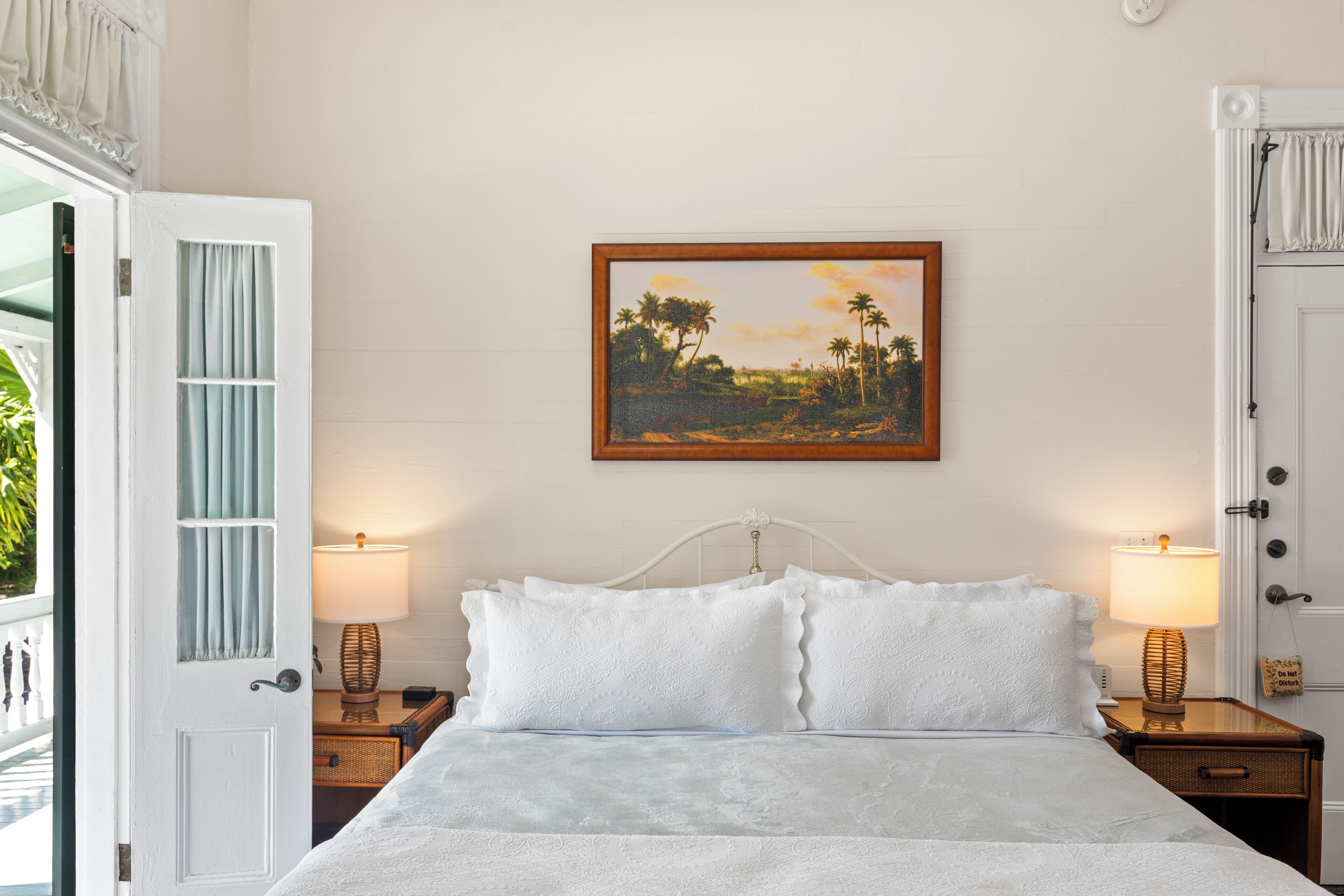 A neatly made bed with white linens, two bedside lamps, a framed painting of a tropical landscape, and a white door with a window.