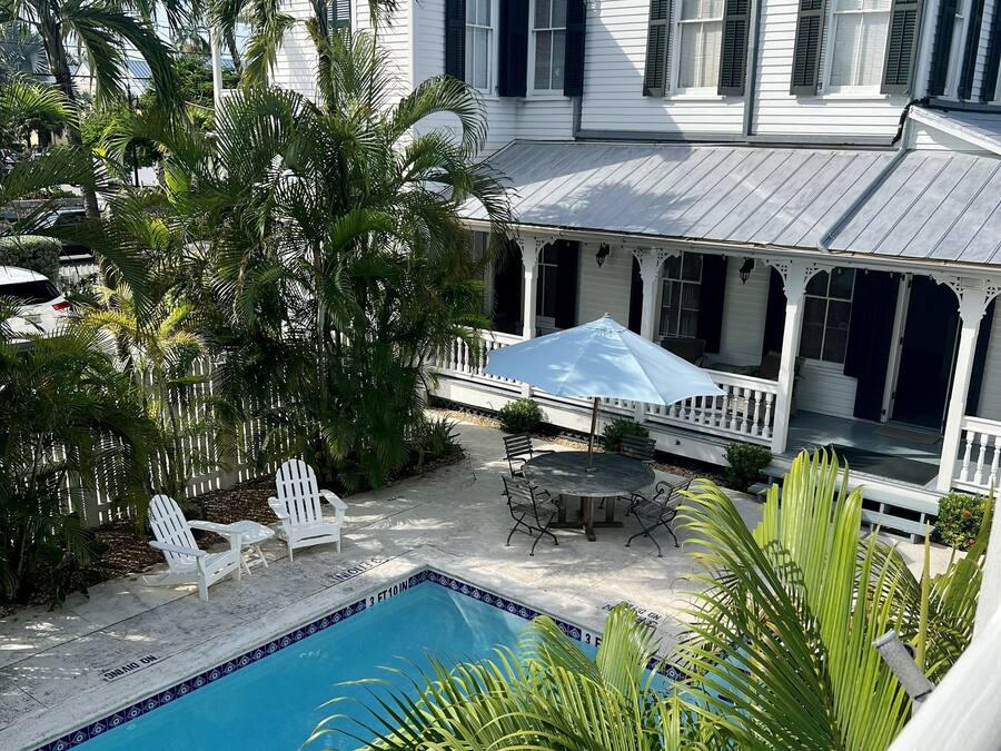 A poolside area with a white house, palm trees, and a covered patio with outdoor furniture.
