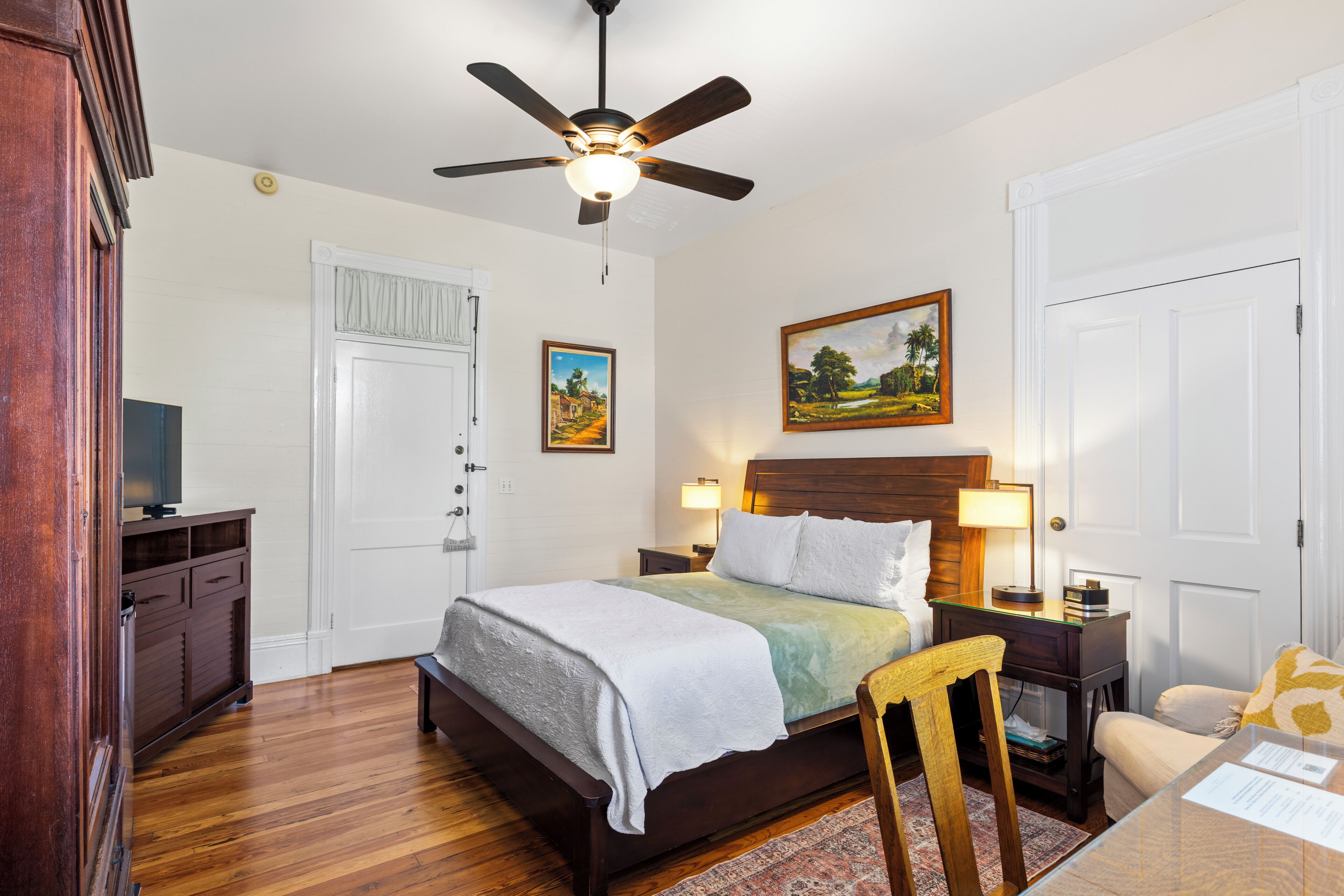 A bedroom with a bed, nightstands, a desk, a chair, a ceiling fan, and a door.