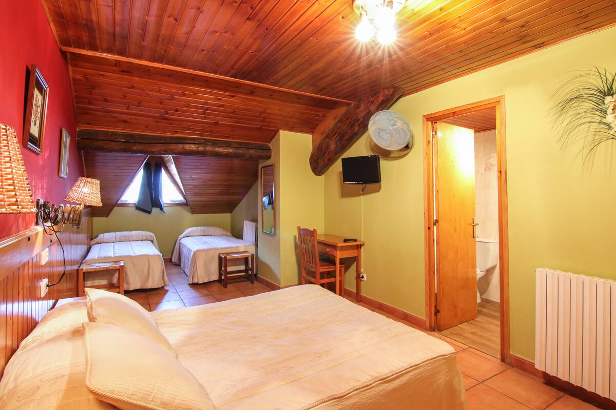 A room with a large bed, two smaller beds, a wooden ceiling, a fan, a TV, and a door leading to a bathroom.