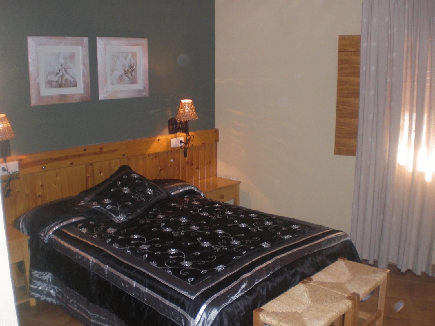 Ein Schlafzimmer mit hölzernem Kopfteil, einer schwarz-blauen Bettdecke mit Blumenmuster, zwei gerahmten Bildern an der Wand und einer Nachttischlampe.