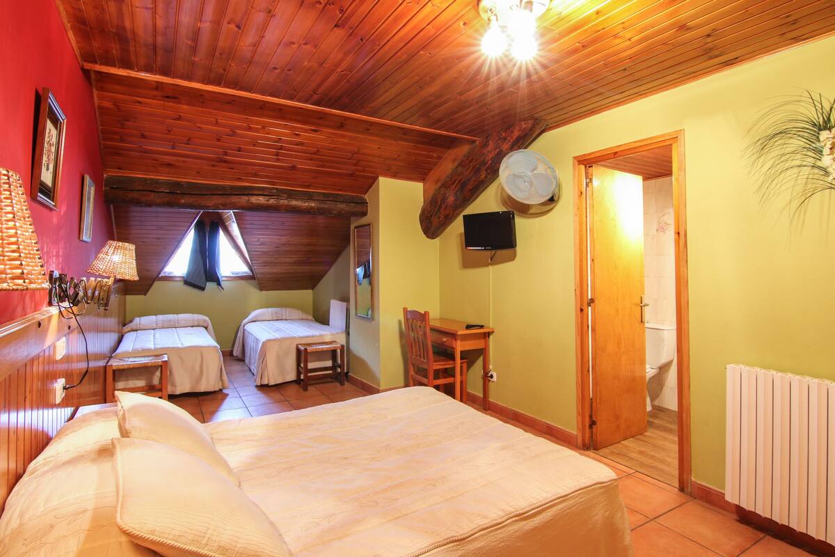 A room with a wooden ceiling, a bed, two smaller beds, a table, a chair, a fan, a TV, and a door leading to a bathroom.