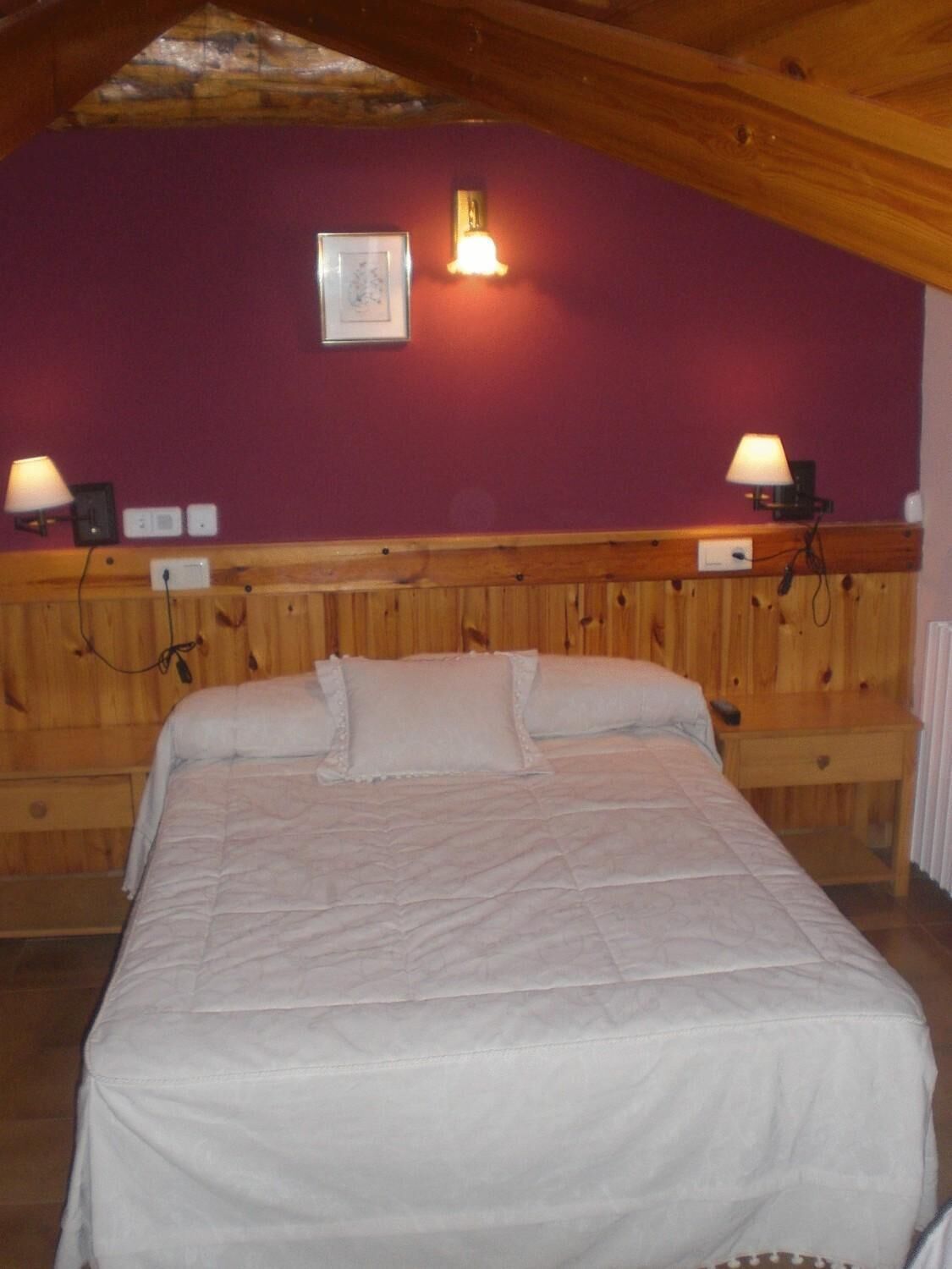 A bedroom with a wooden headboard, a bed with white bedding, two wall-mounted lamps, and a framed picture on the wall.