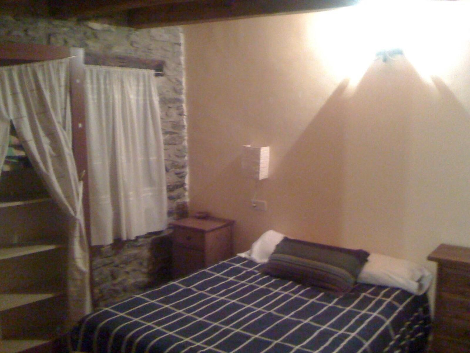 A bedroom with a bed, a nightstand, a lamp, a window with curtains, and a stone wall.