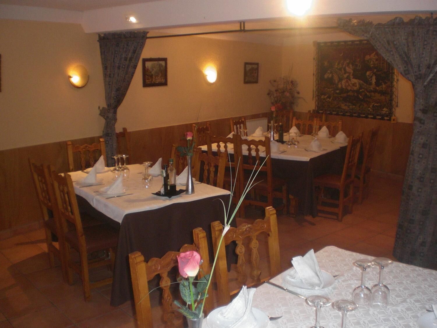 A restaurant with wooden chairs and tables set for dining, featuring a vase with a rose and wine glasses.