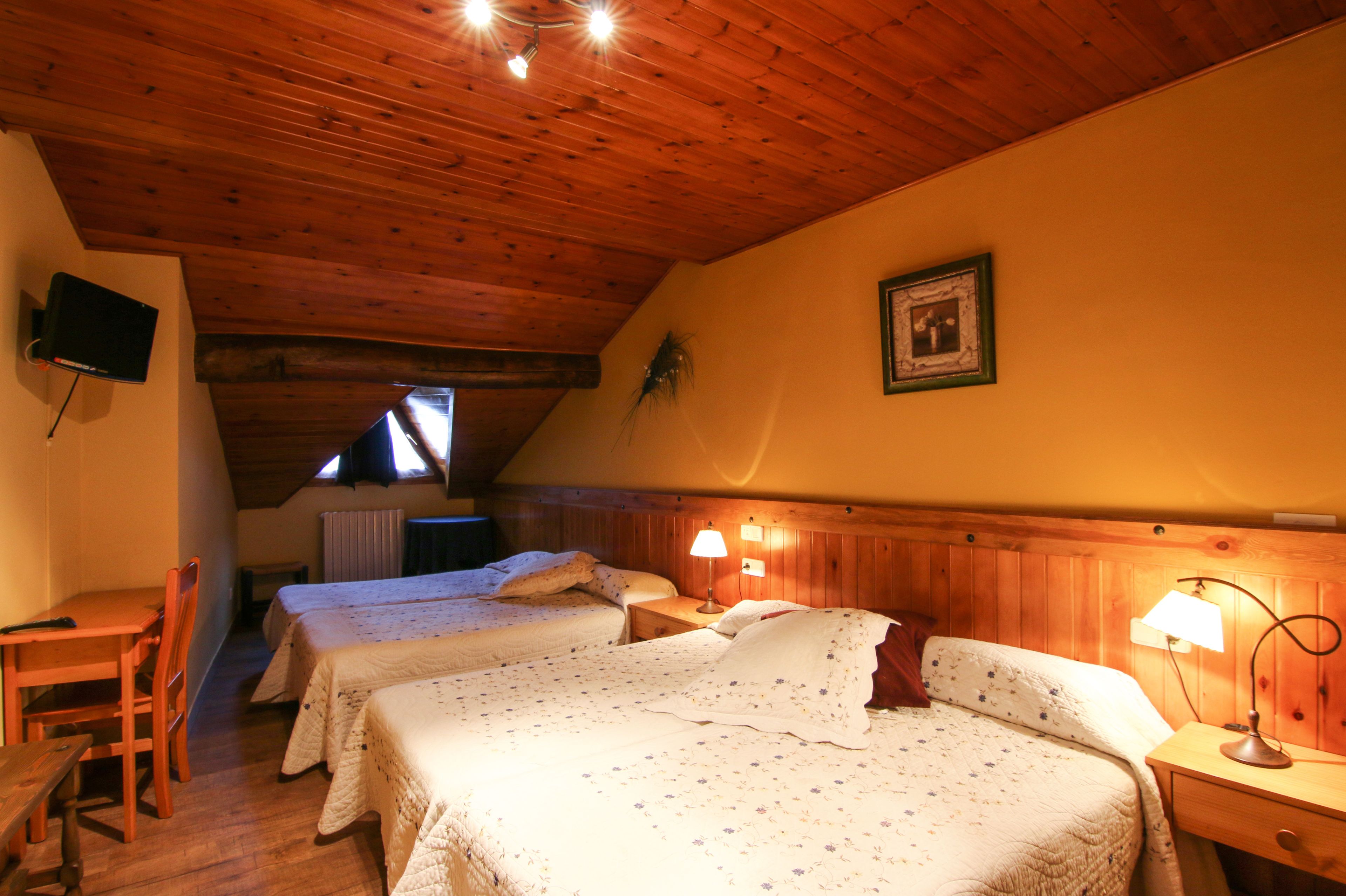 A cozy room with two beds, wooden ceiling, a TV mounted on the wall, and a small desk with a chair.