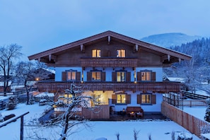 Una casa de madera de dos pisos con un balcón, un paisaje nevado y montañas al fondo.