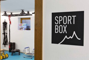 Una habitación con un letrero en la pared que dice 'SPORT BOX', una barra de dominadas, una plataforma escalonada y un estante con diversos objetos.