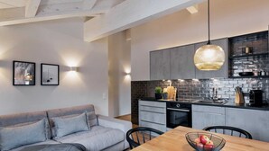 Une cuisine moderne avec un mur de briques noires derrière l’évier, une table à manger en bois sur laquelle est posé un panier de fruits, et un canapé gris dans l’espace salon.