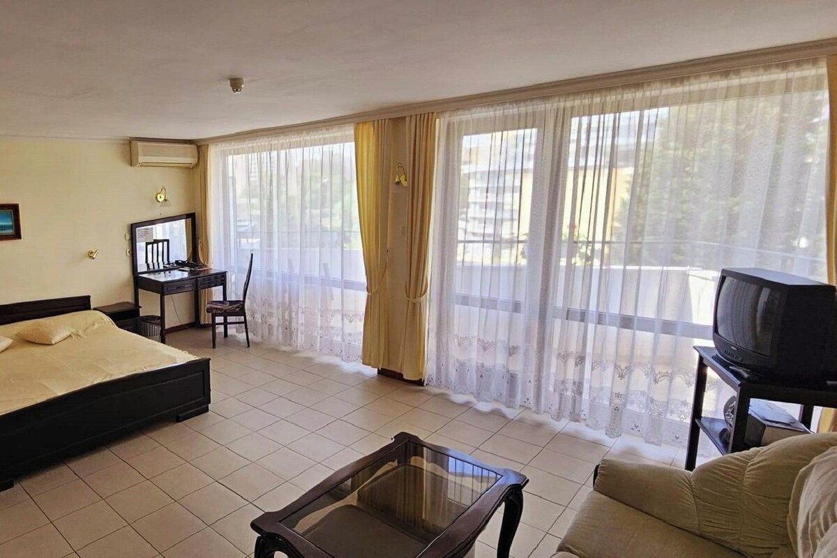 A bedroom with a bed, a desk, a chair, a television, and a balcony with curtains.