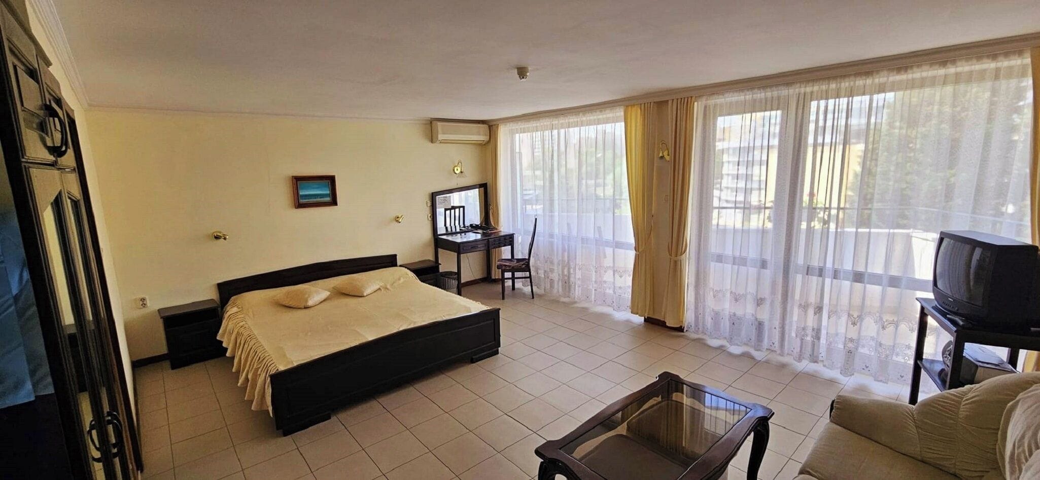 Une chambre à coucher comprenant un lit, un bureau, une chaise, un téléviseur et un balcon avec des rideaux.