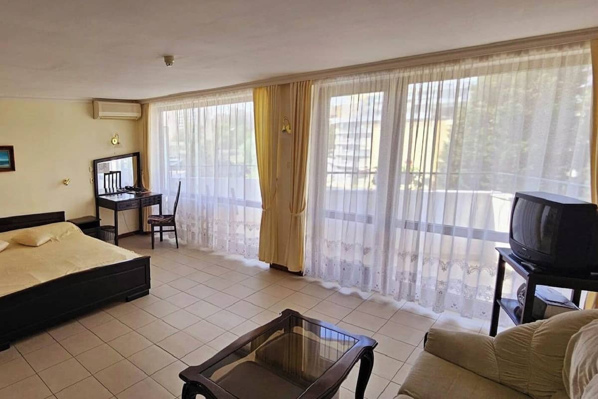 Une chambre à coucher comprenant un lit, un bureau, une chaise, un téléviseur et un balcon avec des rideaux.