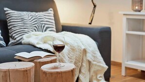 Ein gemütliches Zimmer mit einem grauen Sofa, einem zebra-gemusterten Kissen, einer weißen Strickdecke, einem hölzernen Hocker, einem Glas Rotwein, einem Buch, einer Schreibtischlampe und einem Laternenlicht auf einem weißen Regal.