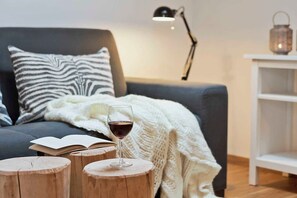 Um quarto acolhedor com um sofá cinzento, uma almofada estampada com padrão de zebra, um cobertor branco de tricô, um banquinho de madeira, um copo de vinho tinto, um livro, uma luminária de mesa e um candeeiro numa prateleira branca.