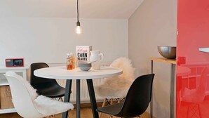 Ein Essbereich mit einem runden Tisch, schwarzen und weißen Stühlen, einer Dose Maisflocken, einer Tasse und einem Radio.
