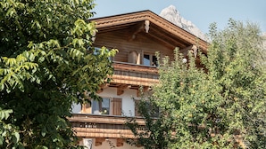 Ein zweistöckiges Holzhaus mit Balkonen, umgeben von Bäumen und einem Berg im Hintergrund.