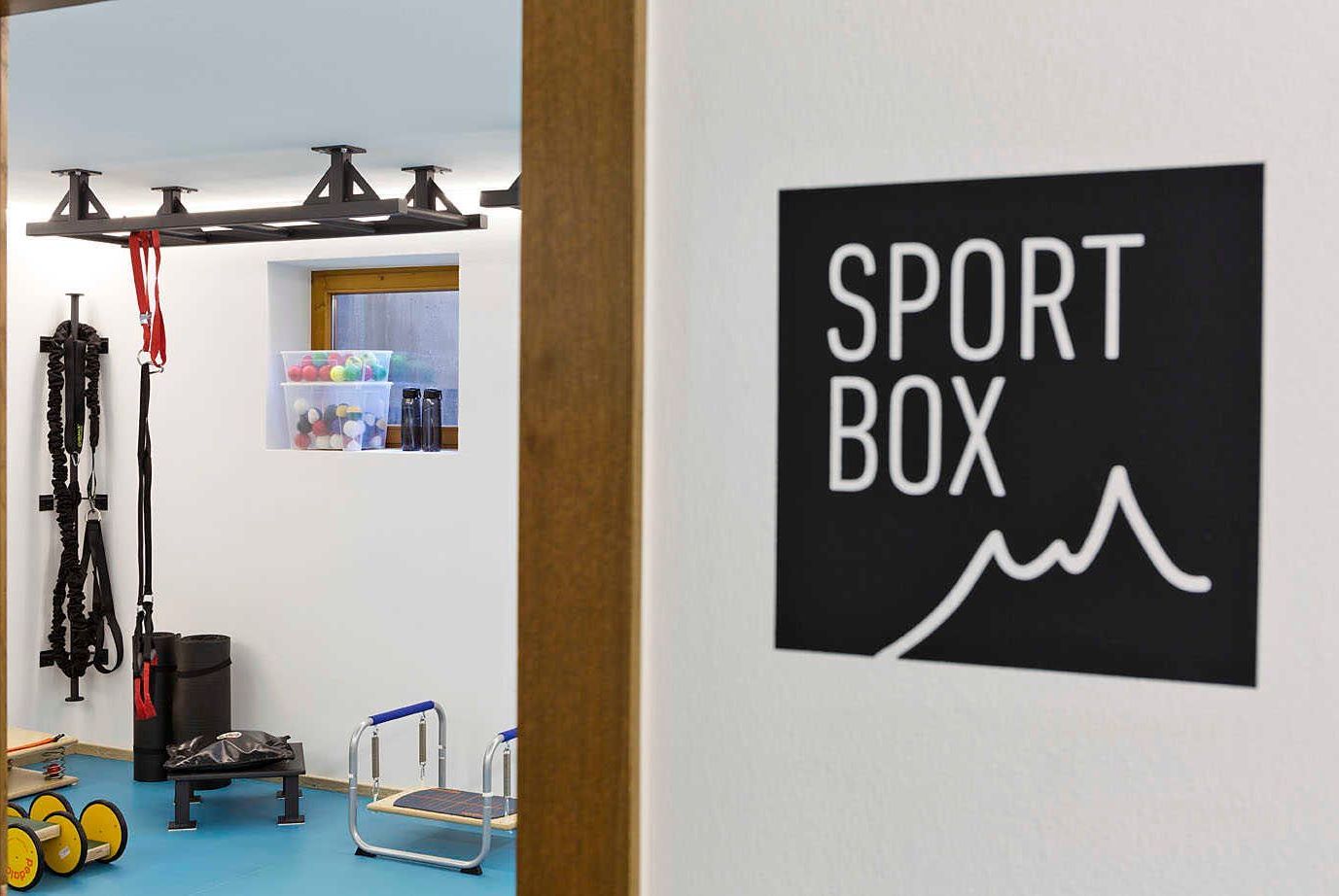 Ein Zimmer mit einer an der Wand befestigten Beschilderung mit dem Schriftzug 'SPORT BOX', einer Klimmzugstange, einer Stehplattform und einem Regal mit diversen Gegenständen.