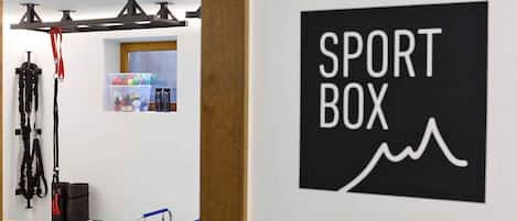Ein Zimmer mit einer an der Wand befestigten Beschilderung mit dem Schriftzug 'SPORT BOX', einer Klimmzugstange, einer Stehplattform und einem Regal mit diversen Gegenständen.