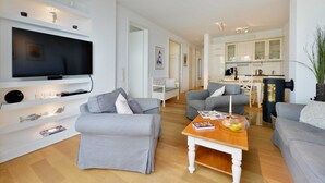 A modern living room with a flat-screen TV, grey sofas, a wooden coffee table, and a kitchen in the background. - Herring Gull, Germany (Sassnitz)