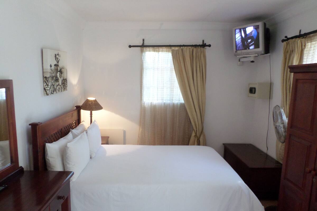 A hotel room with a bed, a bedside table, a lamp, a television on the wall, and a fan.