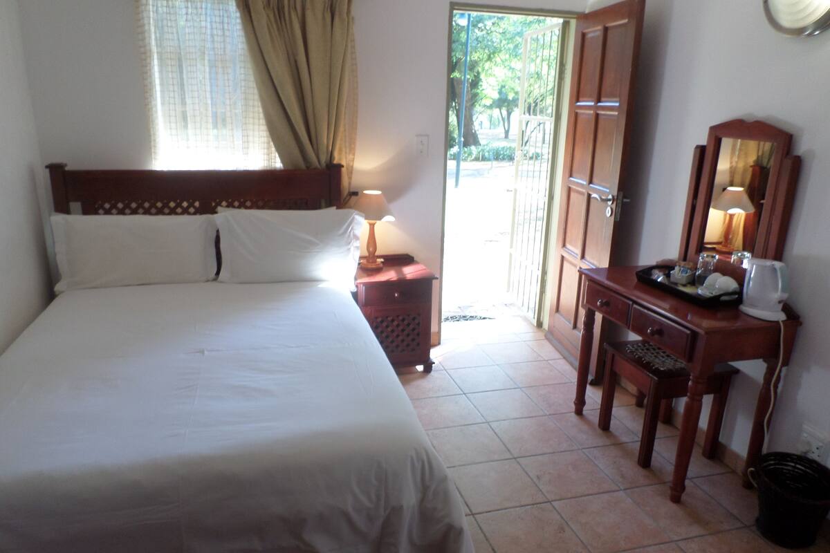 A neatly made bed with white linens, a wooden nightstand with a lamp, a wooden desk with a mirror, and a door leading outside.