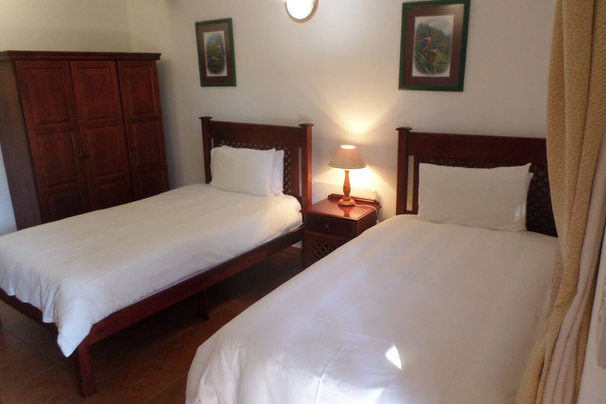 A room with two single beds, wooden headboards, a nightstand with a lamp, and a wardrobe.