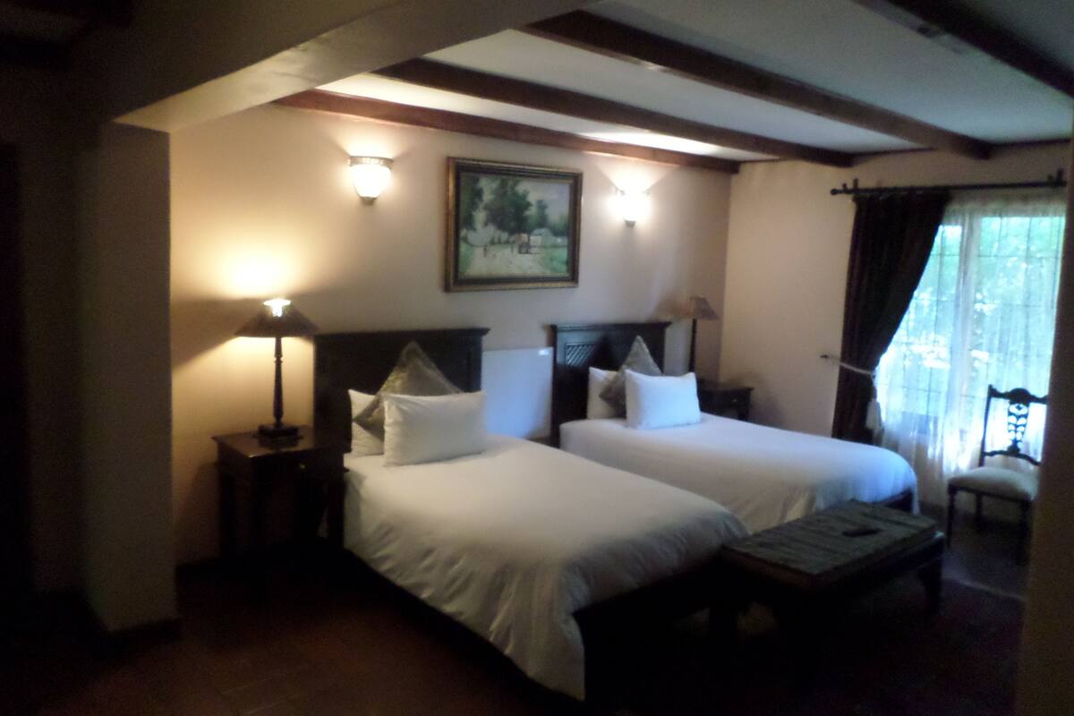 A hotel room with two beds, a nightstand, a lamp, a painting on the wall, and a window with curtains.