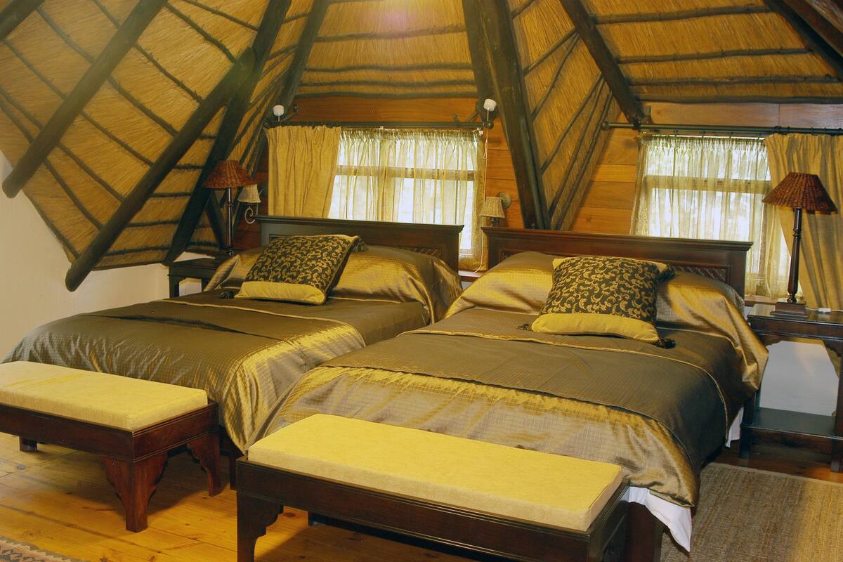 A room with two beds, wooden headboards, a bench, and a thatched roof.