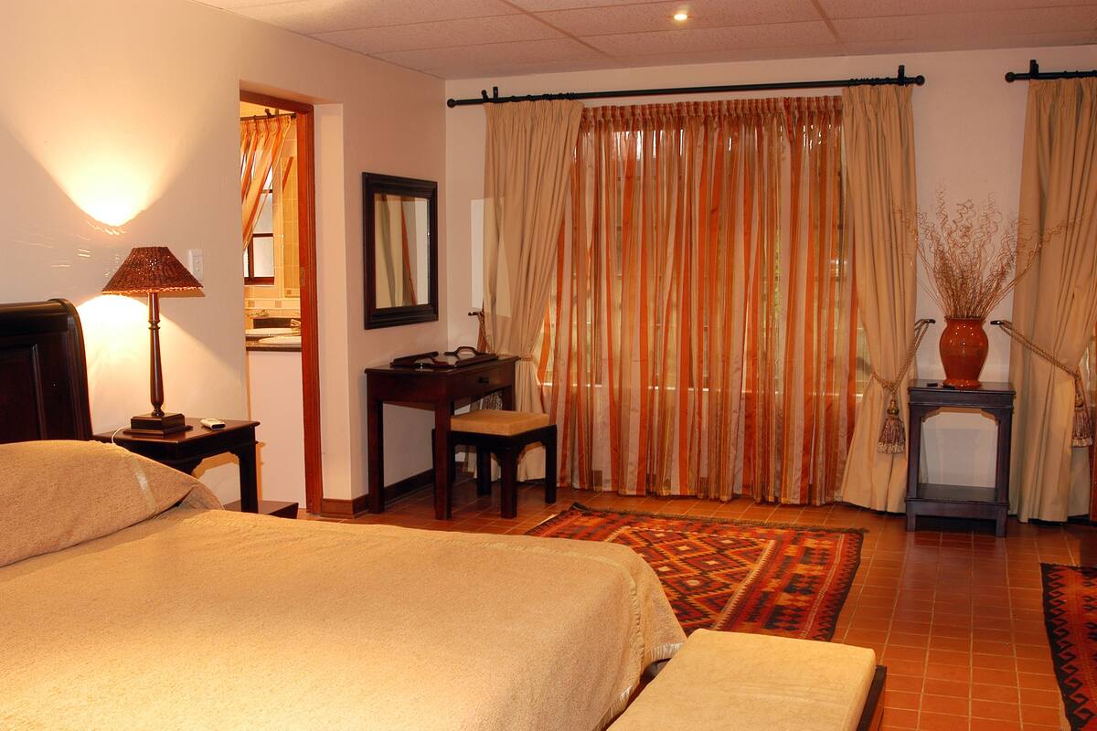 A hotel room with a bed, a desk, a chair, a lamp, a vase with plants, and curtains.
