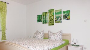 Une chambre à coucher avec un lit, des oreillers verts, une table de chevet avec une lampe et un mur orné de quatre tableaux encadrés.