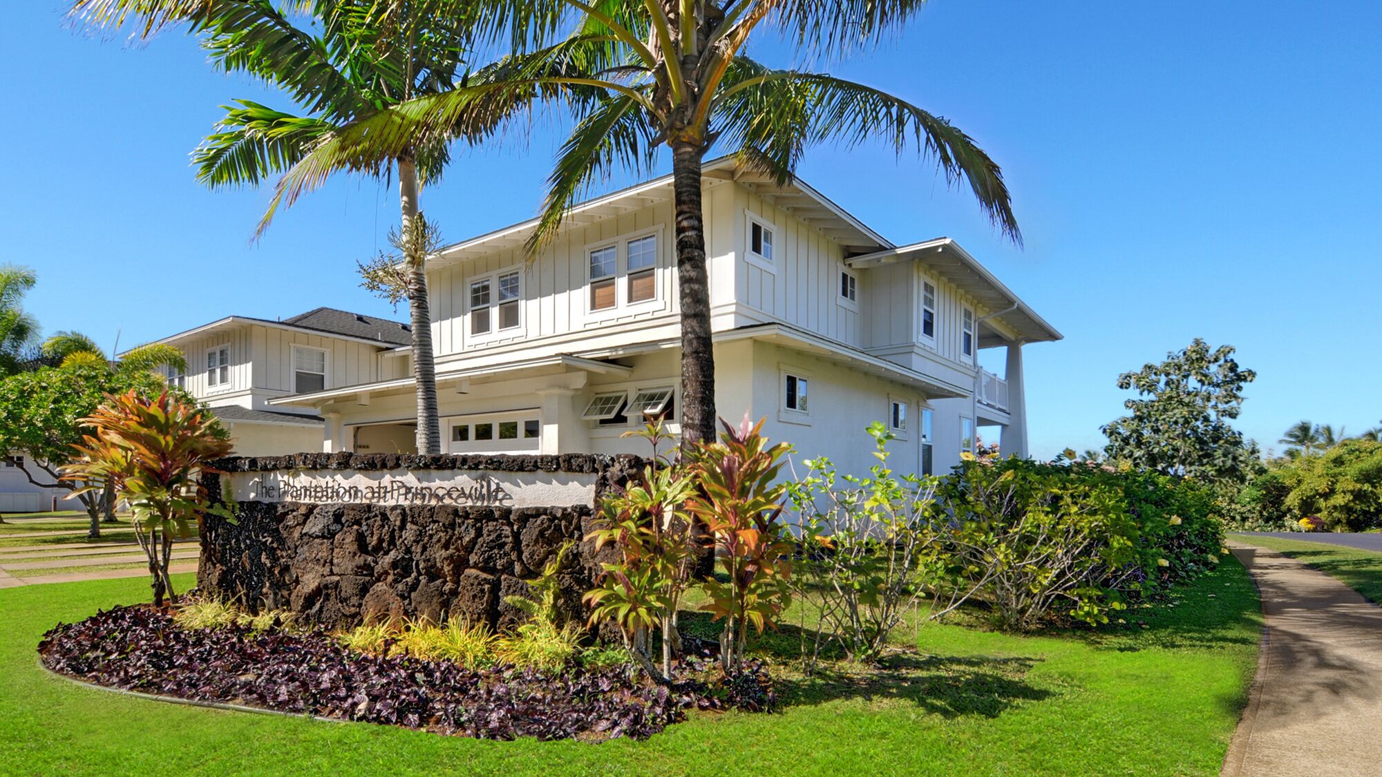A two-story house with a stone wall in front, surrounded by well-maintained grass and tropical plants.