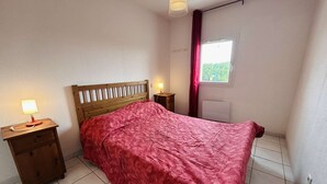 Quarto com cama de madeira, mesa de cabeceira com abajur, janela com vista para vegetação e radiador.