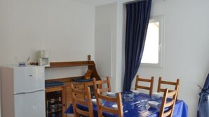 Uma área de refeições compacta com mesa posta para quatro pessoas, cadeiras de madeira, uma pequena geladeira e uma janela com cortinas.