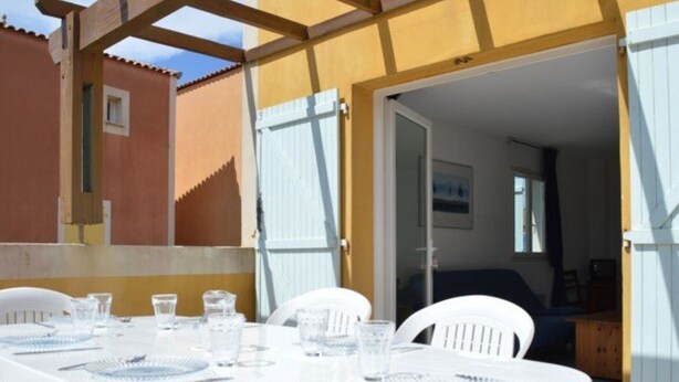 Mesa branca posta para quatro com copos e pratos, uma pérgula de madeira e um edifício amarelo ao fundo.