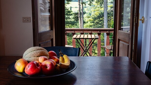 Una mesa de madera con un plato de fruta y vistas a árboles a través de una puerta abierta.