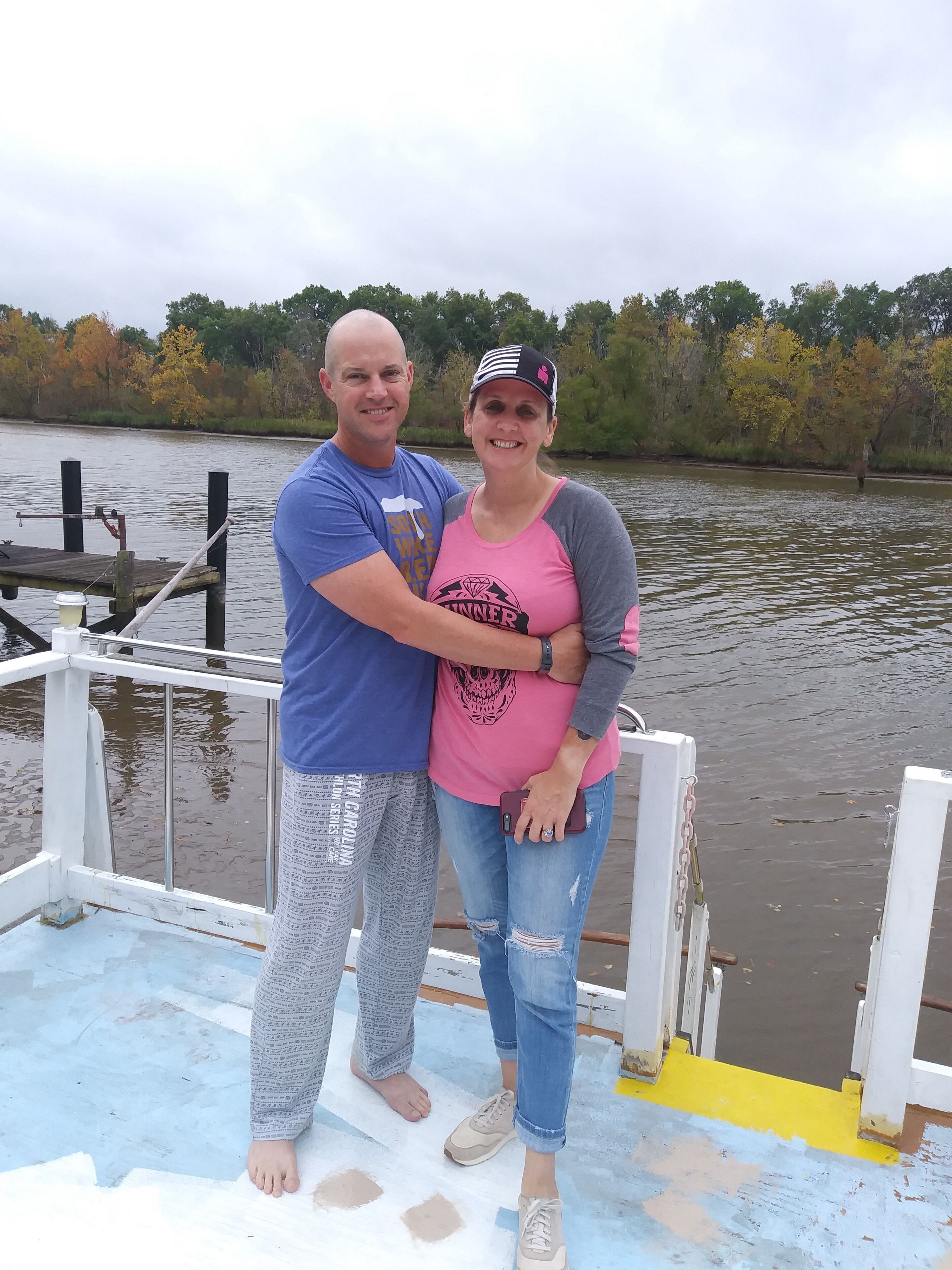 Twee personen staan op het dek van een boot, met een rivier en bomen op de achtergrond.