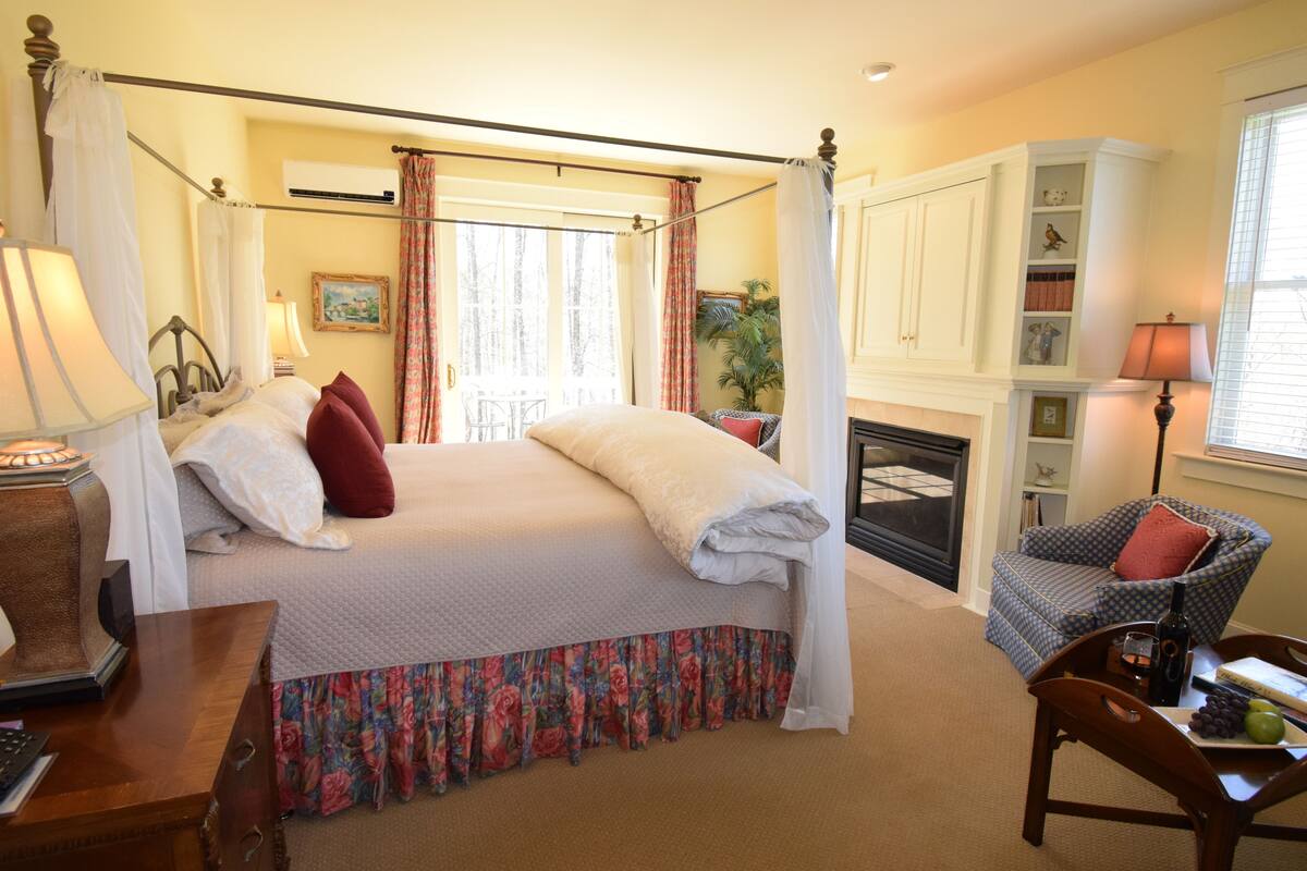 A four-poster bed with a floral bed skirt, a chair with a cushion, a table with a bottle and fruit, and a fireplace.