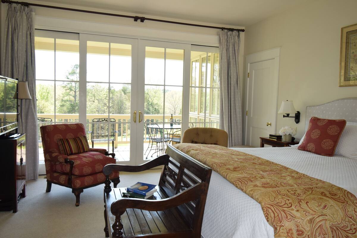 A hotel room with a large bed, a red armchair, a wooden bench, a small table, and a balcony with a chair and table.